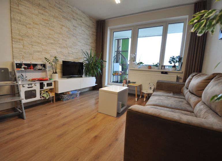 3-IZBOVÝ TEHLOVÝ BYT V DUBNICI NAD VÁHOM | 68 m² | BALKÓN | VLASTNÉ KÚ - Obývačka v 3-izbovom byte s podlahou s dreveným dekorom a kamenným obkladom na stene.