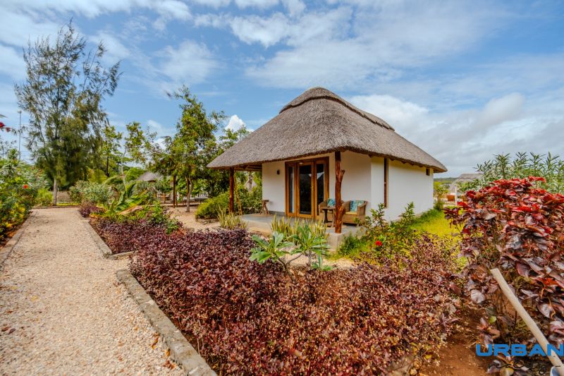 ZARIA GARDEN BUNGALOWS - Rodinný dom so slamenou strechou, obklopený kvetmi a zeleňou, s dláždeným chodníkom.