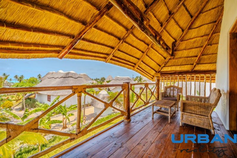 ZARIA GARDEN BUNGALOWS - Drevená terasa s prútenými kreslami, výhľad na tropickú prírodu, výstavba domov v pozadí.