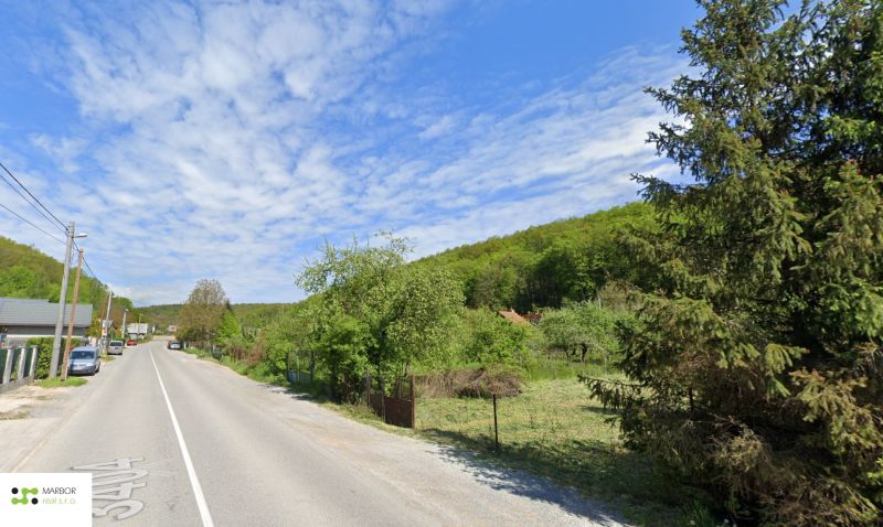 Stavebné pozemky s projektom 2 samostatných RD v obci Baška-časť Maša - Cesta s pozemkami a bývaním, lemovaná stromami, vedúca ku kopcom, obloha s bielymi oblakmi.