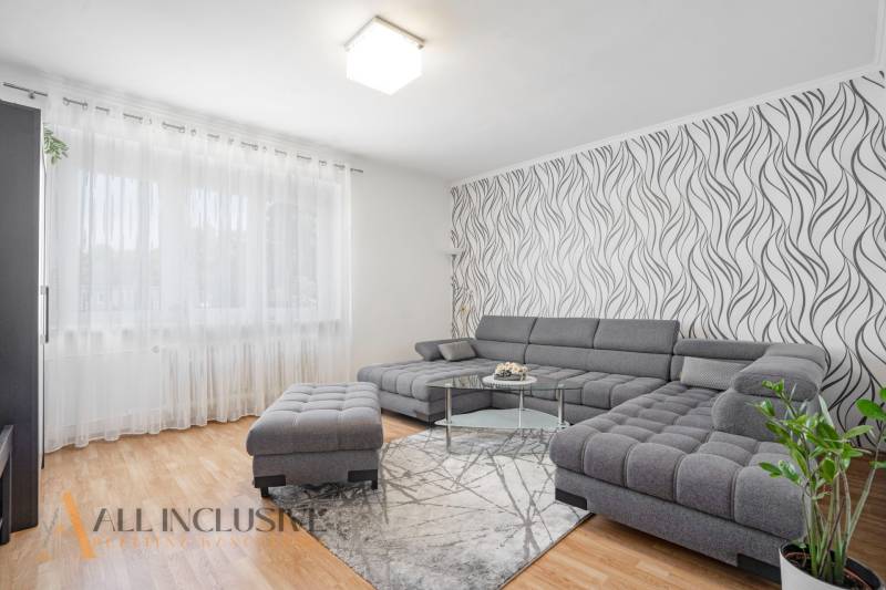 NA PREDAJ ZREKONŠ. 2-IZBOVÝ BYT S BALKÓNOM, GABČÍKOVO - Obývačka 2-izbového bytu s pohodlnou sivou sedačkou, vzorovanou tapetou a drevenou podlahou.