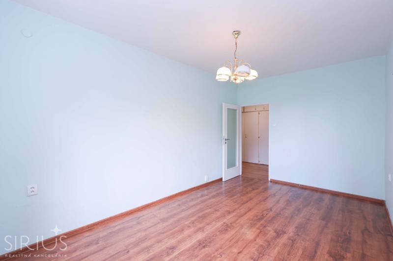 BÝVANIE NA HRADOM KOPCI - 3i byt, 90 m2 s balkónom, SVETLÁ ulica - Prázdna izba v 3-izbovom byte s drevenou podlahou a stropným svietidlom.