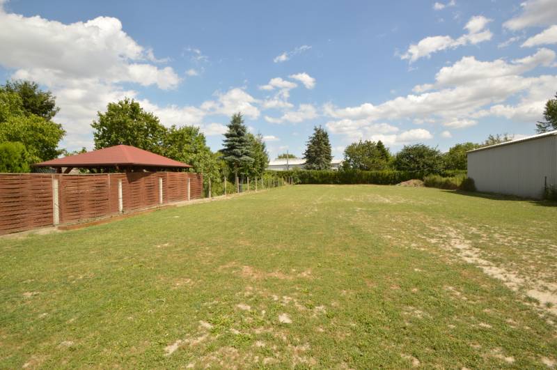 Predaj priestranný 5i s 1483 m2 pozemkom, pivnica, garáž, Dunakiliti - Záhrada pri rodinnom dome v Rajke, tráva, altánok, plot, stromy pod modrou oblohou.