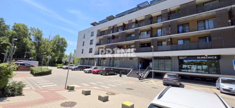 Pekný 2i byt, NOVOSTAVBA, BALKÓN PARKING, KLÍMA, Žltá ulica, SLNEČNICE - Budova so zaparkovanými autami a obchodnými priestormi na Žltej ulici v Petržalke.