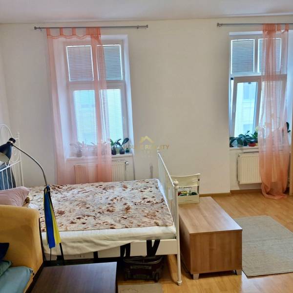 Predaj 1 izbový byt, Karpatská ulica, Bratislava I. Staré Mesto - Interiér 1-izbového bytu s posteľou, drevenou podlahou a oknami s ružovými závesmi.