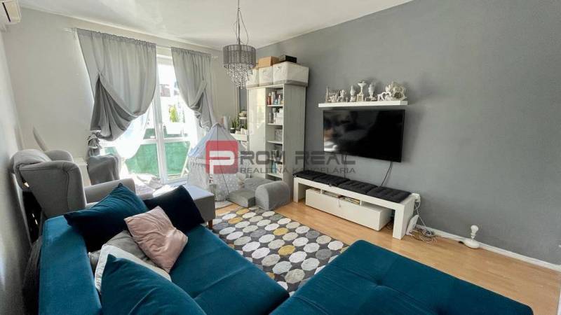 Na PREDAJ priestranný 3 izbový byt s balkónom v meste Senec - Obývačka v 3-izbovom byte s modrou sedacou súpravou a televíziou na stene.