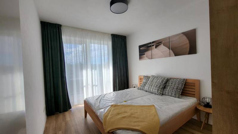 Nový priestranný 3iz. byt s klimatizáciou, balkónom a parkovaním. - Spálňa v 3-izbovom byte s posteľou, obrazmi a vysokými závesmi.