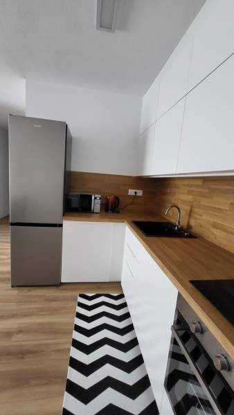Nový priestranný 3iz. byt s klimatizáciou, balkónom a parkovaním. - Kuchyňa v 3-izbovom byte s bielymi skrinkami, drevenou pracovnou doskou a čierno-bielym kobercom.