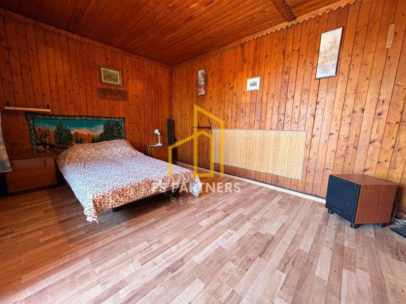 Záhrada s čiastočne prerobenou chatkou – Komárno, časť Nová Stráž - Drevený interiér so posteľou, dekoratívna tkanina na stene, obrazy a jednoduchý nábytok.
