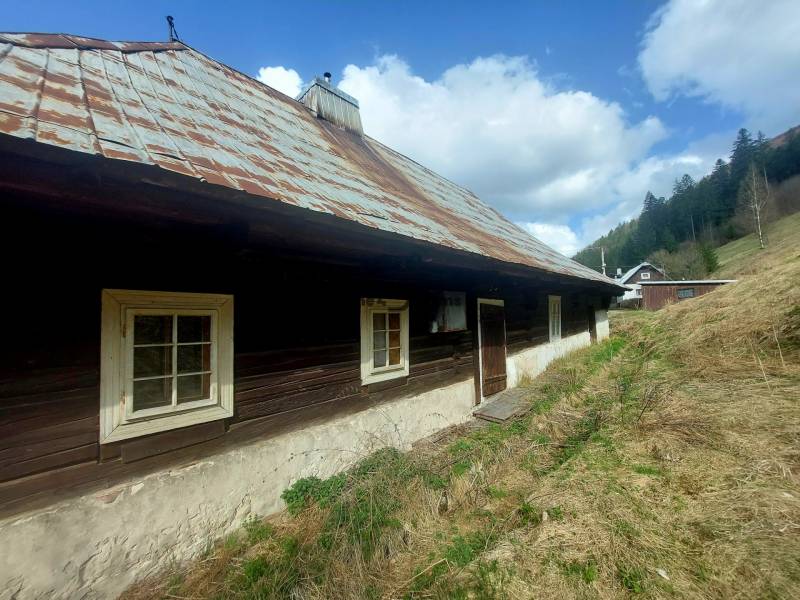 NA PREDAJ drevená chalúpka v Motyčkách  - Drevená chalupa s plechovou strechou na Motyčkách, obklopená prírodou a oblohou plnou oblakov.