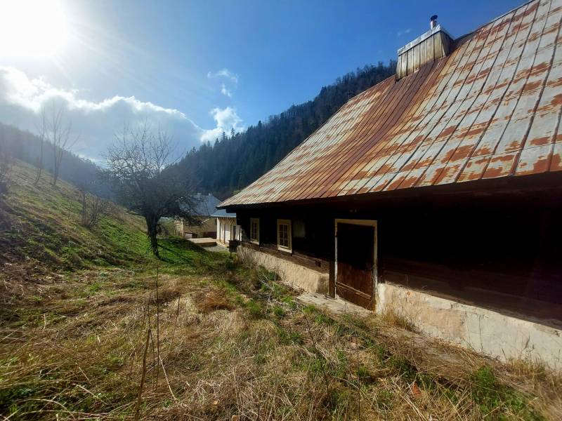NA PREDAJ drevená chalúpka v Motyčkách  - Drevená chata so starou strechou v prírode pod horami, slnečný deň.