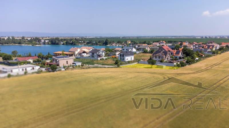  Stavebný pozemok pri jazere Nové Košariská  - Pozemky - bývanie v Dunajskej Lužnej s výhľadom na jazero, domy a pole.