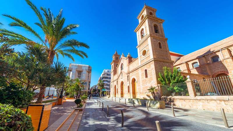TORREVIEJA - UVÁDZACIE CENY ZA NOVOSTAVBU len 300 metrov od mora - Ulica Campoamor 12 v Torrevieji s kostolom a palmami za slnečného dňa.
