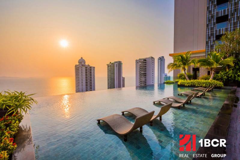 THE RIVIERA WONGAMAT BEACH PATTAYA - Bazén, ležadlá a panoráma výstavby bytov v Pattaye pri západe slnka.
