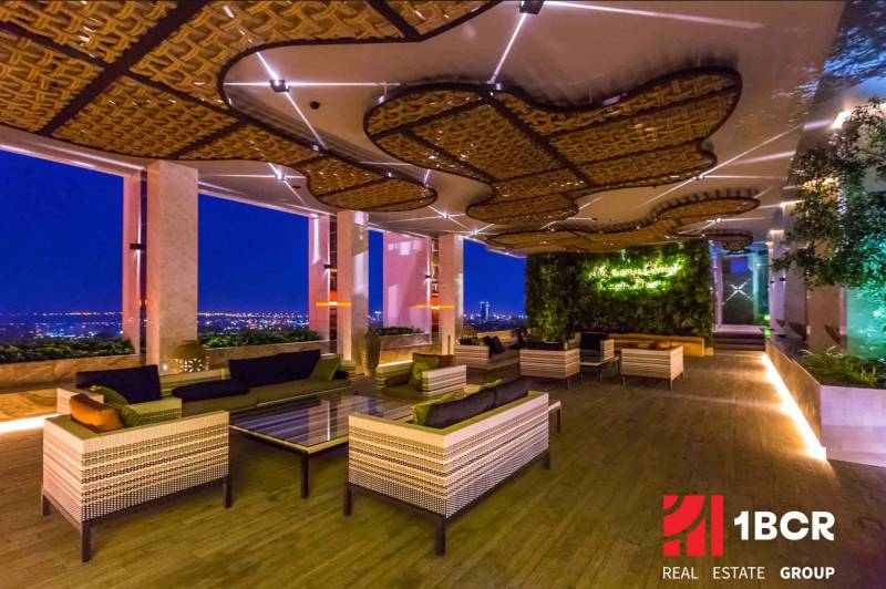 THE RIVIERA WONGAMAT BEACH PATTAYA - Luxusný bytový priestor s výhľadom na večerné osvetlenie mesta, zelenou stenou a pohodlným sedením.