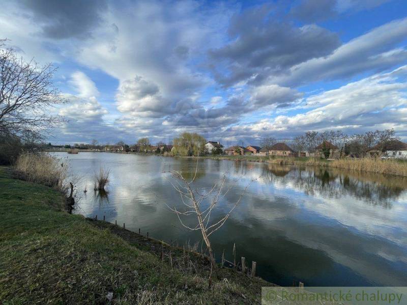 Priestranný pozemok so starším rodinným domom v obci Tvrdošovce - Idylická krajina s jazerom, oblakmi a rodinnými domami v pozadí pri brehu.