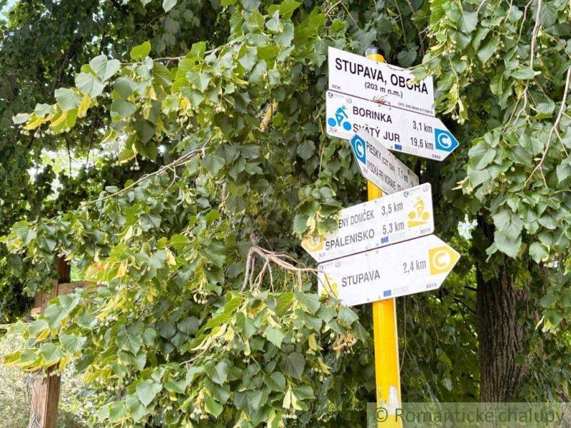 Dve susediace záhrady –12 árov v Stupave, s potenciálom na rekreáciu a - Turistické smerovníky obklopené lístím v Záhradách Malaciek, ukazujúce turistické trasy.