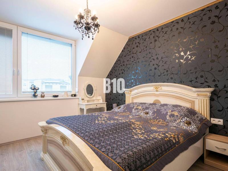 Predaj rodinného domu, Malacky - Spálňa v rodinnom dome s dekoratívnou tapetou, elegantným lusterom a oknami so žalúziami.