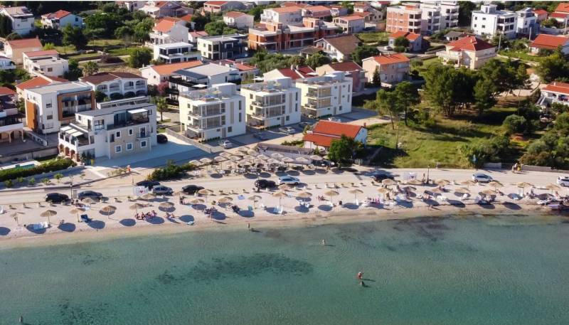 CHORVÁTSKO - Luxusné apartmány (4i) - ostrov VIR - Pláž s lehátkami a slnečníkmi, za ňou domy a budovy v letovisku.