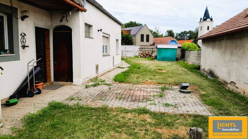 Predaj rodinného domu, Neusiedl am See - Dvor rodinného domu v Potzneusiedli s trávnikom, záhradným domčekom a kostolnou vežou v pozadí.