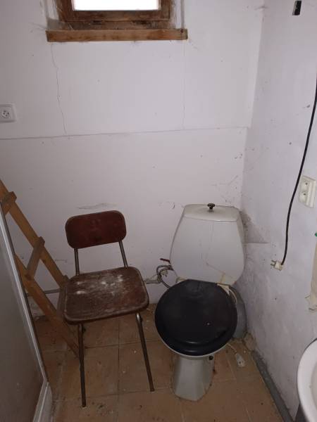 Na predaj rekreačná chata s pozemkom – Nová Baňa (záhradkárska osada) - Záchod na chate s dreveným oknom, stoličkou a popraskanými stenami.