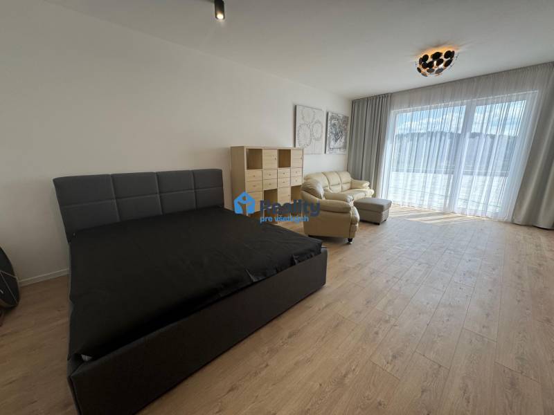 Prenájom NOVOSTAVBA FIM moderný byt 50 m2 + terasa 53 m2 PB - Izba v 2-izbovom byte s posteľou, pohovkou a veľkým oknom s bielymi závesmi.