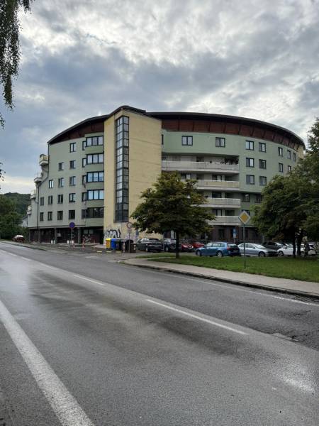 Garsónka v tehlovom dome. Dúbravka - Bytový dom v Bratislave - Dúbravke, obklopený ulicou, stromami a zaparkovanými autami.