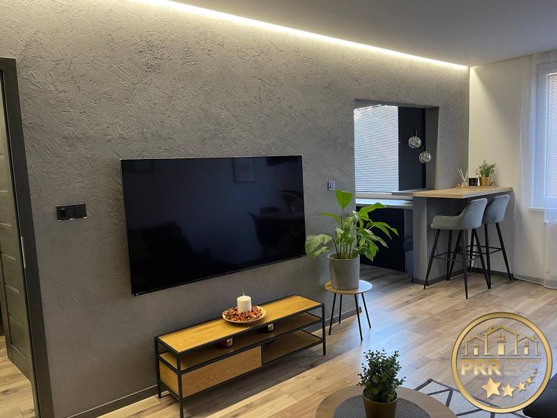 Na predaj krásny komplet prerobený, moderne zariadený 3-izb. byt v BnB - Obývačka v 3-izbovom byte s moderným TV, rastlinami a barovým pultom s vysokými stoličkami.