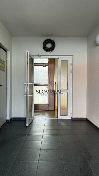 ✅ SLOVREAL ponúka na predaj Garsónku v obľúbenej lokalite Levice – Ryb - Vchod do budovy s dlažbou, dverami s presklením, veniec na stene a jasné svetlo.