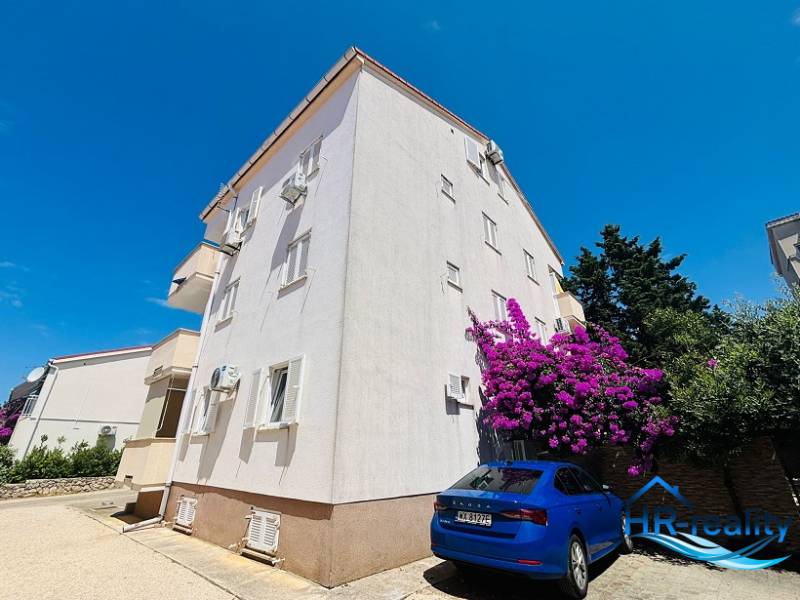 ☀NOVALJA (HR) -  2-izbový apartmán 45 m2 s terasou v 2.rade u mora - Bytový dom v Novalji s parkovaním, bielo-kvetnaté stromy a modré auto pred budovou.
