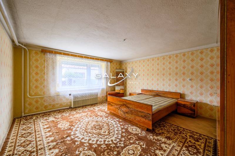 Rodinný dom s veľkým pozemkom na predaj – Vidiná, okres Lučenec - Spálňa s posteľou, vzorovanými tapetami a kobercami v rodinnom dome.