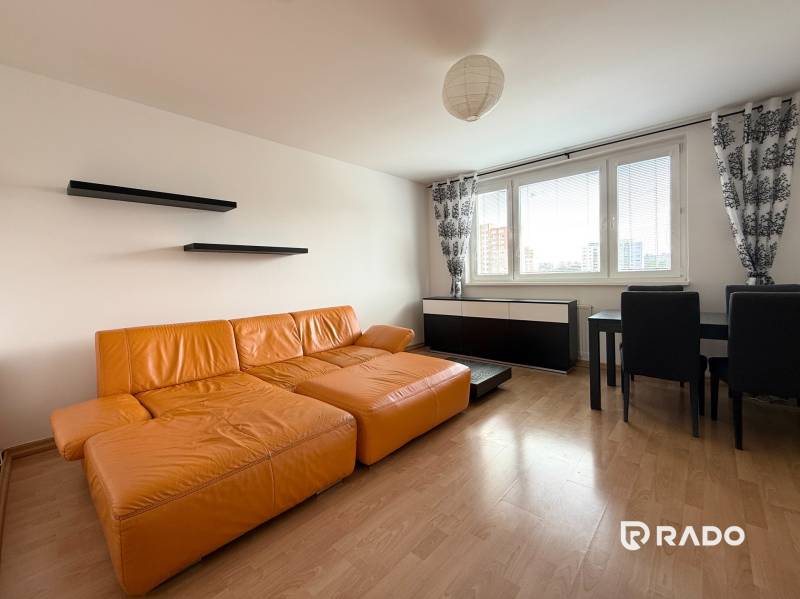 Priestranný 3-izbový byt s balkónom 68 m² Belinského - Obývačka v 3-izbovom byte s oranžovou sedačkou, čiernymi policami a jedálenským stolom.