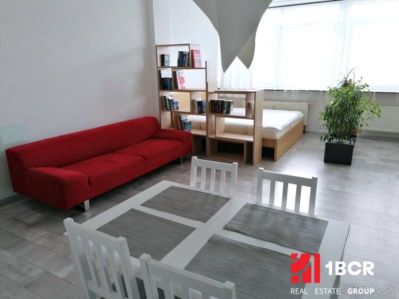 VELKOMETRÁŽNY 1 IZB.AP.BYT- TRNAVSKÁ CESTA, NOVÉ MESTO,BRATISLAVA - Obývací priestor s červenou pohovkou, stolom a posteľou v 1-izbovom byte.
