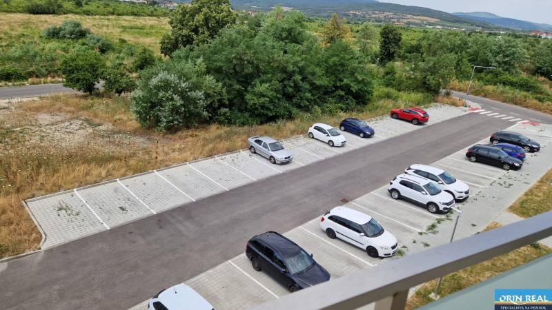 NOVÝ ZARIADENÝ BYT NA Muškáte - Parkovisko pred 2-izbovým bytom na Muškátovej ulici v Pezinku, obklopené zeleňou a kopcami.