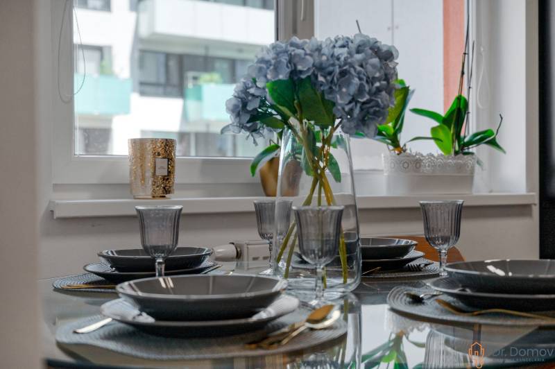 NA PREDAJ EXKLUZÍVNY 2-IZBOVÝ BYT S TERASOU, NOVOSTAVBA, JÉGÉHO ALEJ - Elegantne prestretý stôl s kvetmi v 2-izbovom byte, sklenený riad a príbor.