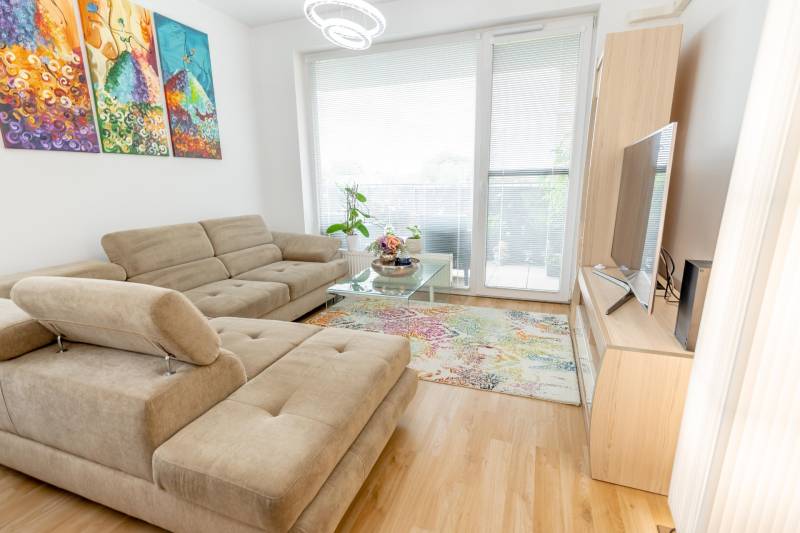 Jedinečný 3-izbový apartmán s balkónom, 71 m², Nový Ružinov - Obývacia izba s pohodlnou pohovkou, farebnými obrazmi a veľkými oknami v 3-izbovom byte.