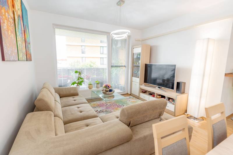 Jedinečný 3-izbový apartmán s balkónom, 71 m², Nový Ružinov - Svetlá obývačka 3-izbového bytu s pohovkou, televízorom, kobercom a kvetmi pri okne.