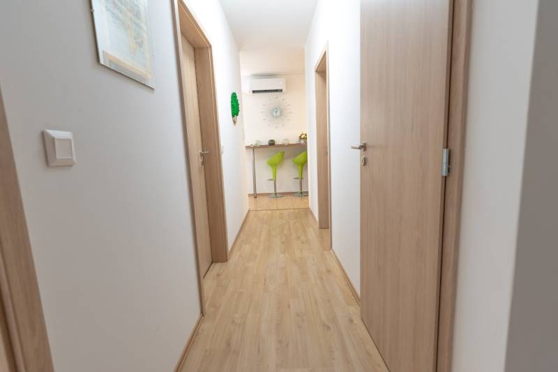 Jedinečný 3-izbový apartmán s balkónom, 71 m², Nový Ružinov - Chodba s drevenou podlahou, vedúca k zeleným barovým stoličkám v 3-izbovom byte.