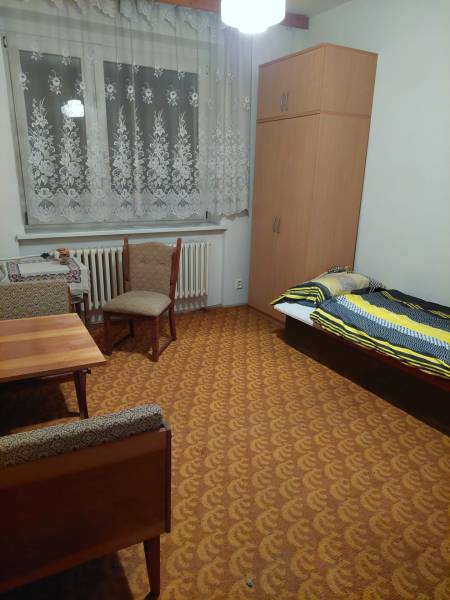 VÝNIMOČNÁ INVESTIČNÁ PRÍLEŽITOSŤ V CENTRE PIEŠŤAN - Izba rodinného domu s posteľou, skriňou, stolom a stoličkami, ladená v teplých farbách.
