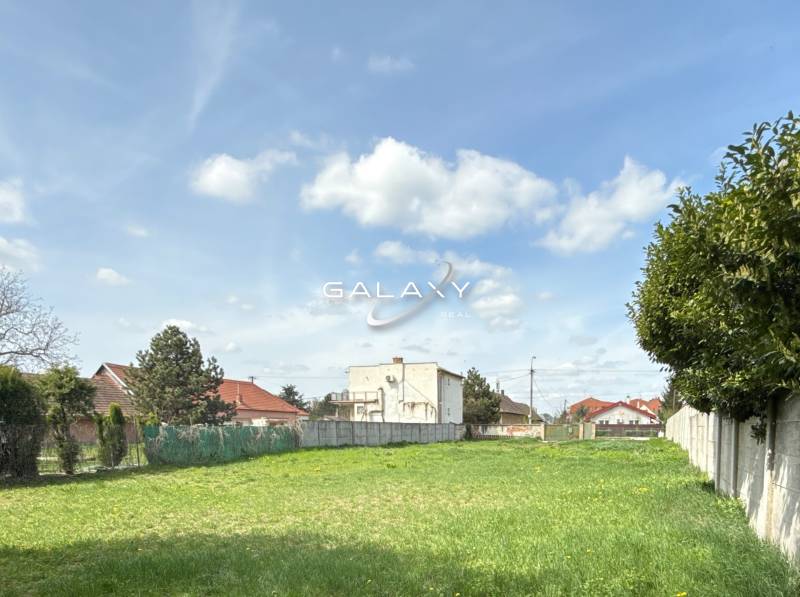 Pozemok 1315 m² pre rodinný dom blízko centra – Sládkovičovo - Pozemok na bývanie v Sládkovičove, tráva, ploty, stromy, budovy v pozadí, oblačná obloha.