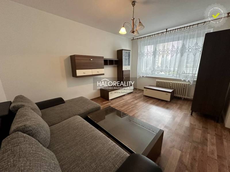 Prenájom, jednoizbový byt Rimavská Sobota, P.Hostins... - 1-izbový byt s pohovkou, dreveným nábytkom a veľkým oknom s čipkovanou záclonou.