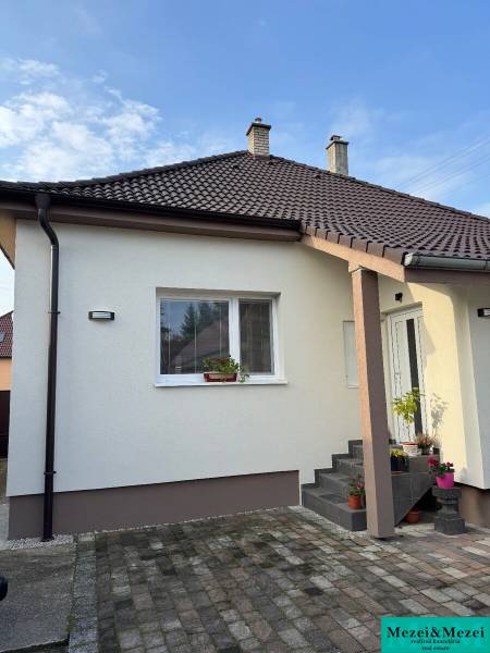 ? Na predaj: Rodinný dom s vedľajšou stavbou – Trstin (okres Trnava) - Rodinný dom v Trstíne s bielou fasádou, hnedou strechou a vydláždeným chodníkom.