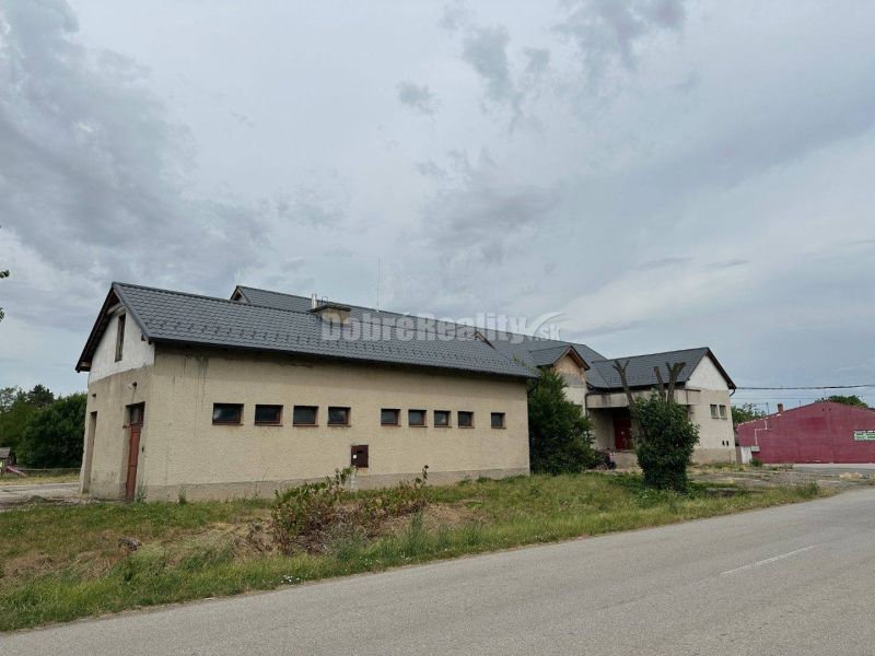 Dobré reality - Polyfunkčný objekt na predaj - Budova v Tekovských Lužanoch s trávnatým okolím pri ceste pod zamračenou oblohou.