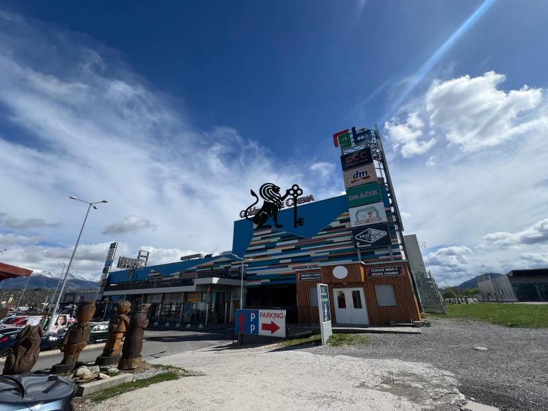 Prenájom obchodných priestorov v nákupnom centre RGB Liptov - Obchodné priestory na ulici Kamenné pole, parkovanie a sochy z dreva pri vchode.