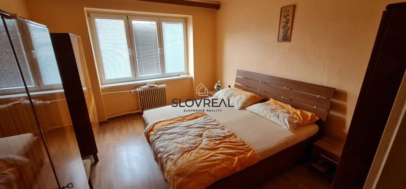SLOVREAL - na predaj 2 izbový byt Hodruša - Hámre, 58 m2 - Spálňa v 2-izbovom byte s dreveným nábytkom, posteľou a veľkým oknom.