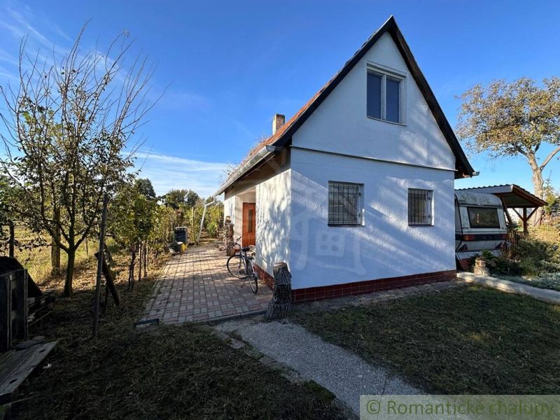 Chatka s rozsiahlou záhradou v lone prírody pri mlynskom náhone v Nový - Chata v Nových Zámkoch s patio, stromami a prívesom pod modrou oblohou.