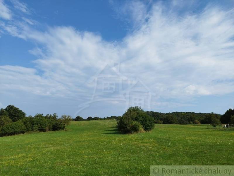 Veľký, vyše 4,5Ha slnečný pozemok s chalupou v strede, na lazoch, pri  - Lúka v Hrušove obklopená lesmi, pod modrou oblohou s oblakmi.