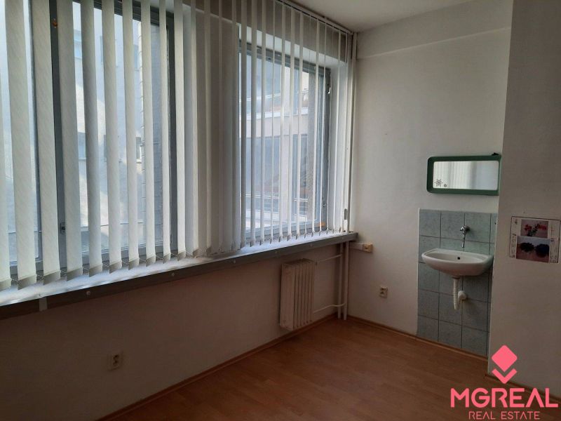 Prenájom kancelárií 21 m2 v centre Trnavy - Kancelária s umývadlom a podlahou s dreveným dekorom, svetlo cez vertikálne žalúzie.