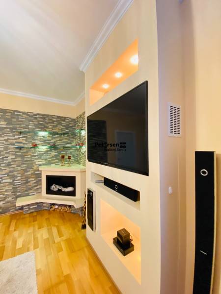 NA PRENÁJOM PRIESTRANNÝ 3-IZBOVÝ BYT V SKVELEJ LOKALITE, UL. SLOBODY - Obývacia stena s televízorom, dekoratívnym kameňom a podlahou s dreveným dekorom v 3-izbovom byte.