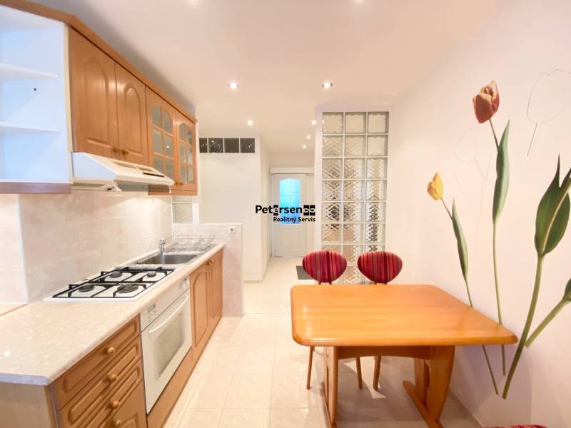 NA PRENÁJOM PRIESTRANNÝ 3-IZBOVÝ BYT V SKVELEJ LOKALITE, UL. SLOBODY - Kuchyňa v 3-izbovom byte s drevenými skrinkami, stolom a dekoratívnou stenou s kvetmi.